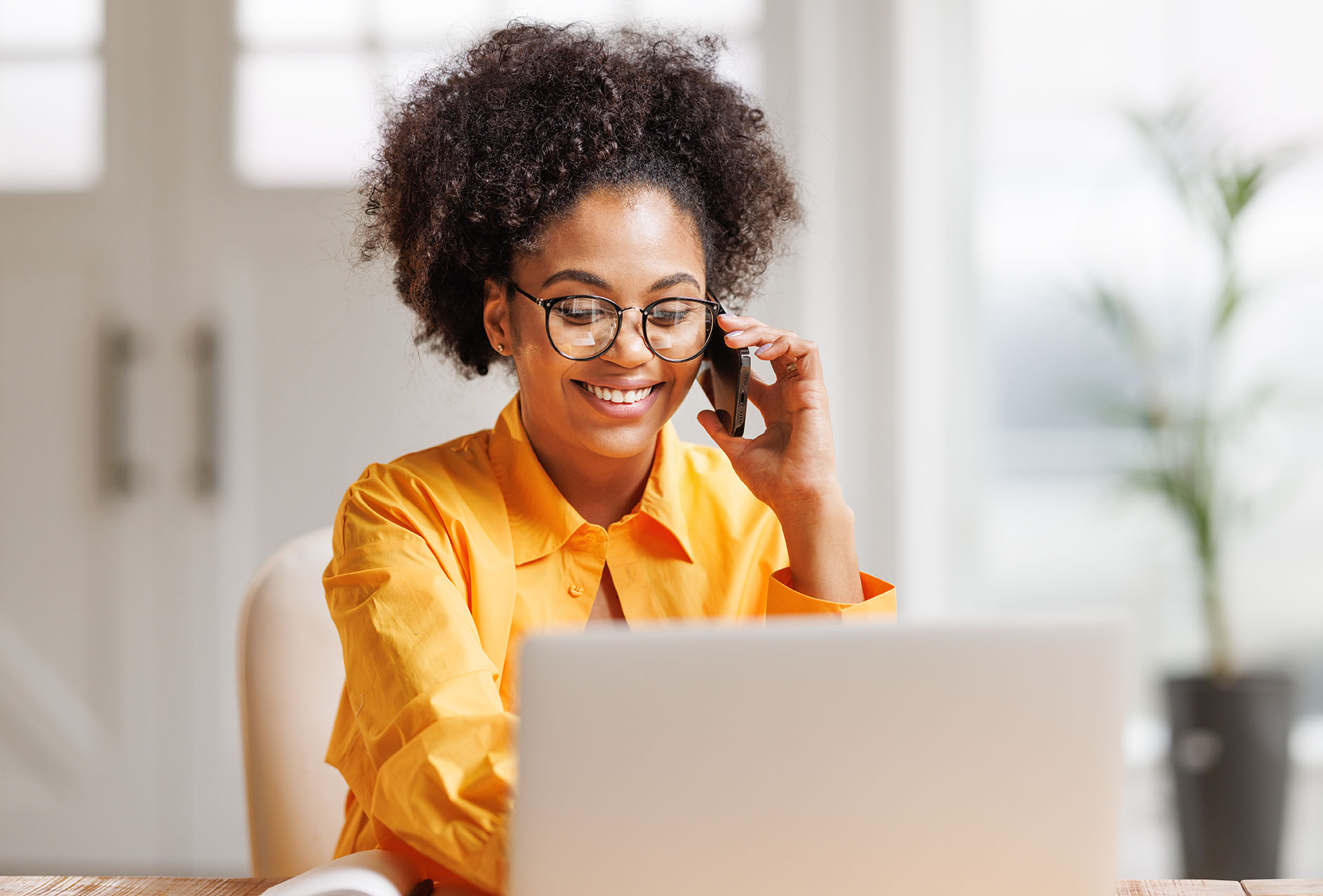 Beautiful young smiling ethnic woman making call via smartphone while working remotely from home while sitting at desk and talking to coworkers